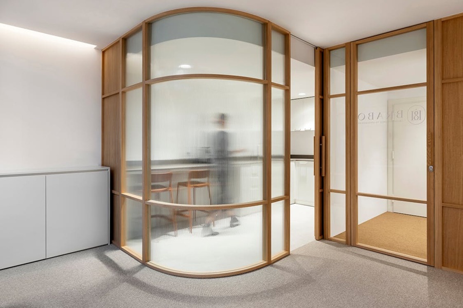 Separador de ambientes curvo con marco de madera y paneles de cristal estriado en oficina de diseño.