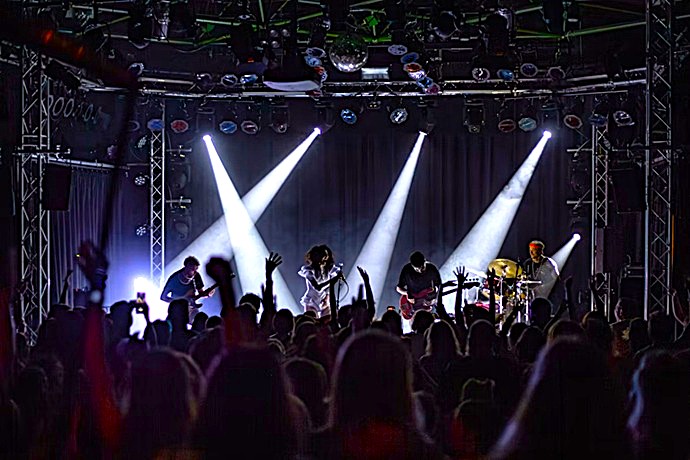 Escenario de concierto con banda en directo y diseño de iluminación basado en potentes haces blancos verticales proyectados desde una estructura técnica elevada.