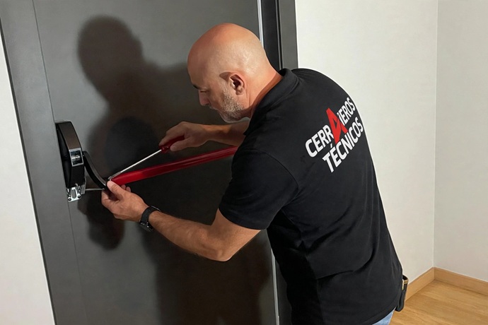 Técnico de Cerrajeros Técnicos ajustando con un destornillador el mecanismo de fijación de una barra antipánico roja en una puerta de emergencia.